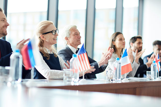 A group of business people sitting at a table clapping with flags from different countries in front - by pressmaster / Adobe Stock A group of business people sitting at a table clapping with flags from different countries in front - by pressmaster / Adobe Stock
